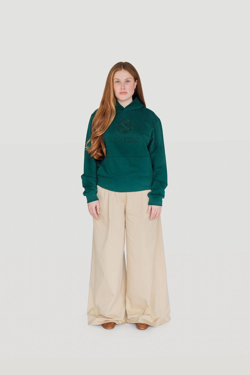 Front view of a woman wearing the green Gratitude hoodie with cream wide-leg trousers