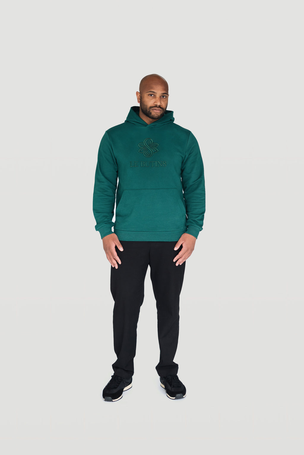 Front view of a man wearing the green Le Butin8 Gratitude hoodie with black trousers