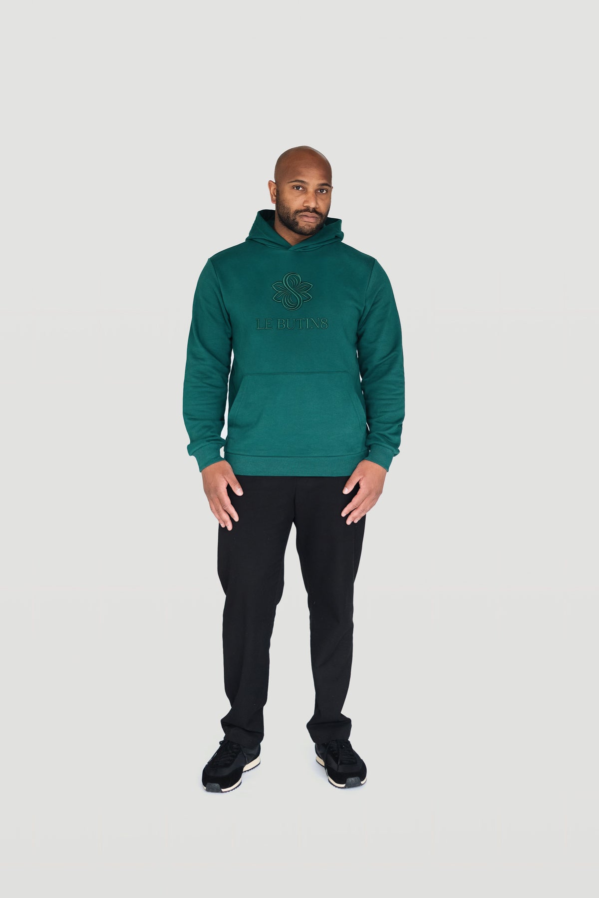 Front view of a man wearing the green Le Butin8 Gratitude hoodie with black trousers