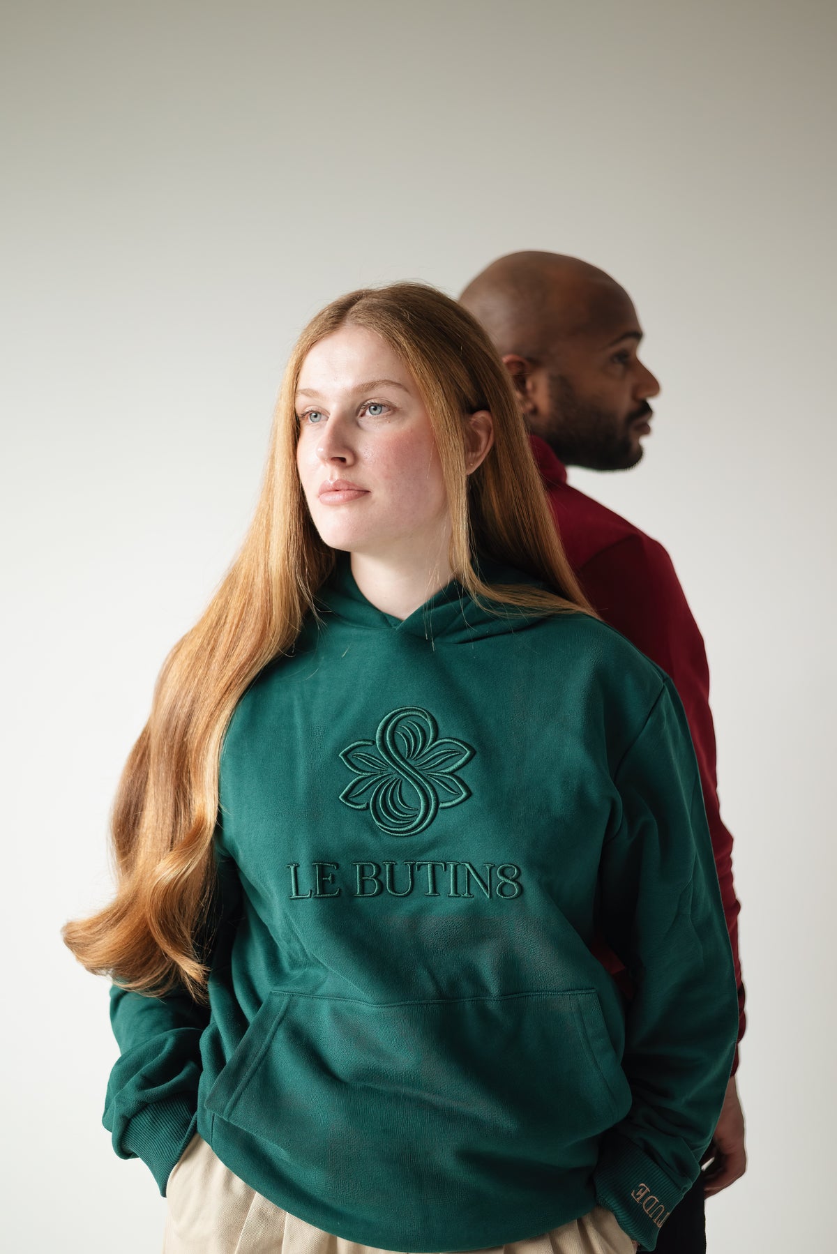 Woman wearing the green Le Butin8 Gratitude hoodie with a model in bordeaux behind her