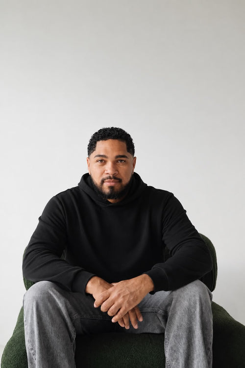 Man seated wearing the black Le Butin8 Believe hoodie with grey trousers